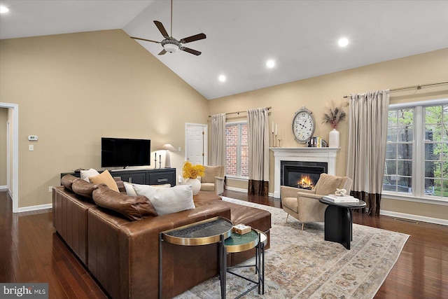 living area with high vaulted ceiling, a glass covered fireplace, ceiling fan, baseboards, and dark wood-style flooring