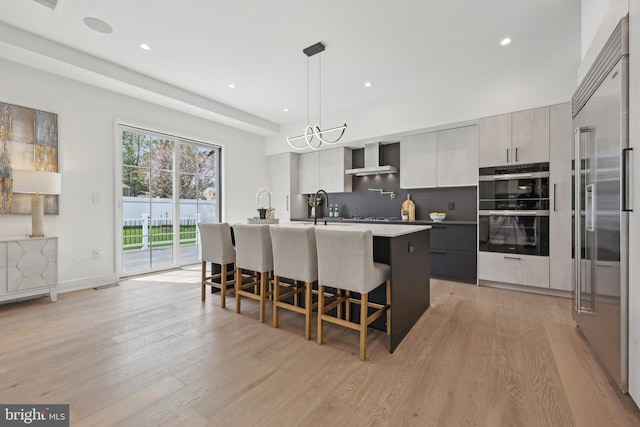 kitchen featuring modern cabinets, stainless steel built in refrigerator, multiple ovens, and wall chimney exhaust hood