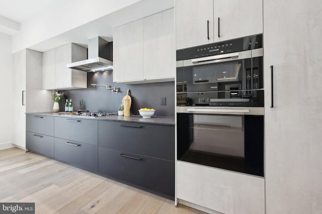 kitchen featuring multiple ovens, stainless steel gas stovetop, light wood-type flooring, modern cabinets, and wall chimney exhaust hood
