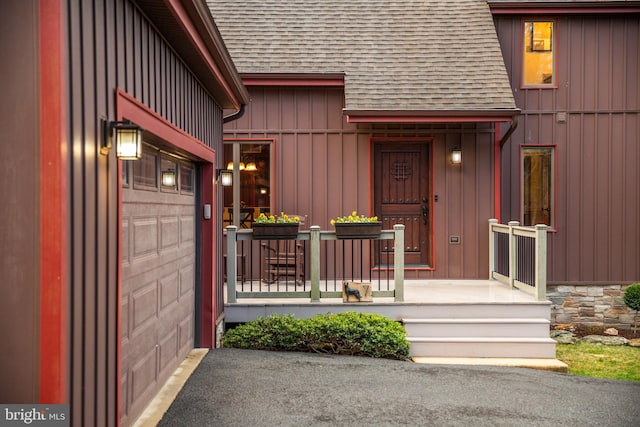 view of exterior entry with board and batten siding, a garage, a shingled roof, and aphalt driveway