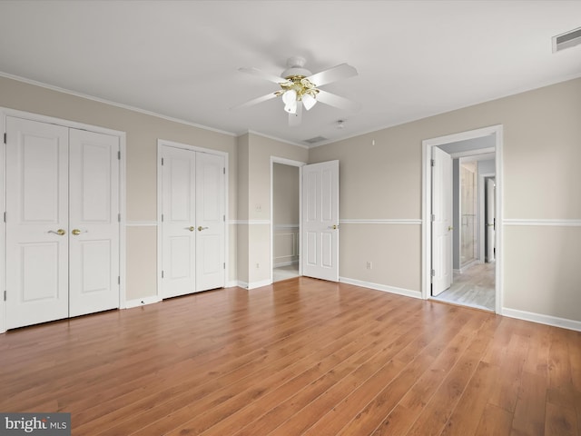 unfurnished bedroom featuring crown molding, two closets, light wood-style flooring, baseboards, and visible vents