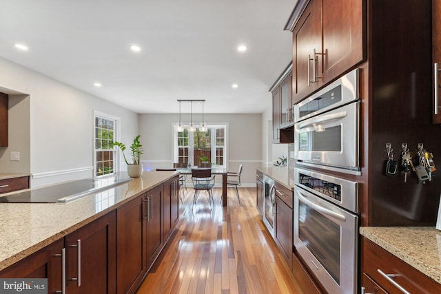kitchen featuring oven, black electric cooktop, light wood-style floors, hanging light fixtures, and light stone counters