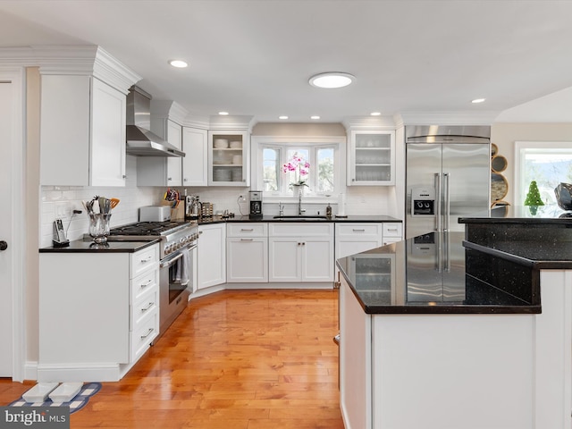 kitchen with high end appliances, light wood-type flooring, wall chimney range hood, white cabinetry, and a sink