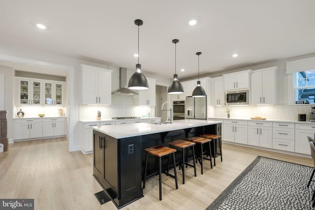 kitchen with light wood-type flooring, wall chimney range hood, white cabinets, a center island with sink, and appliances with stainless steel finishes