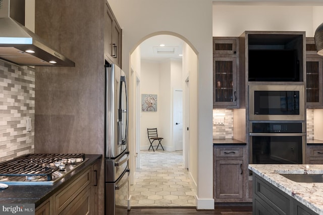 kitchen with wall chimney range hood, dark stone countertops, decorative backsplash, arched walkways, and stainless steel appliances