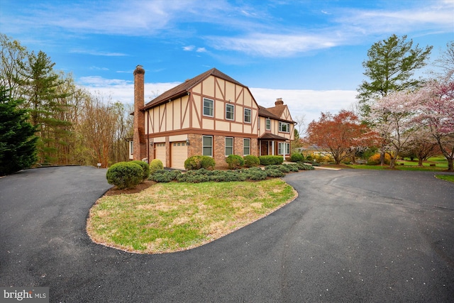 tudor home with an attached garage, brick siding, driveway, and a chimney