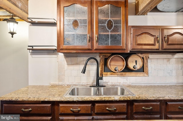 kitchen with tasteful backsplash, dark brown cabinets, a sink, glass insert cabinets, and light stone countertops