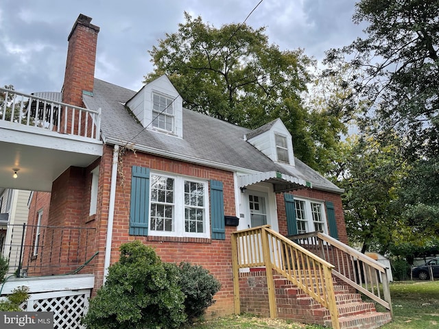 view of front facade with brick siding, a balcony, a chimney, and a shingled roof
