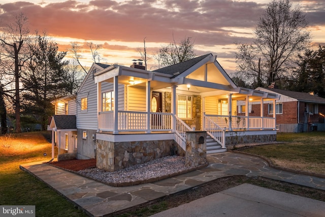 view of front facade with a porch, stone siding, a front lawn, a chimney, and a shingled roof