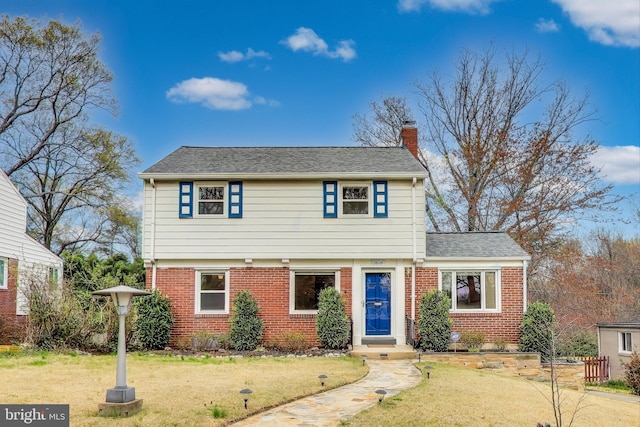 colonial home featuring brick siding, a front yard, roof with shingles, and a chimney