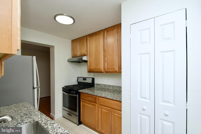 kitchen featuring stainless steel range with gas stovetop, freestanding refrigerator, under cabinet range hood, light tile patterned flooring, and light stone counters