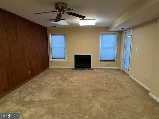 unfurnished living room featuring a fireplace with flush hearth, baseboards, carpet floors, wooden walls, and ceiling fan