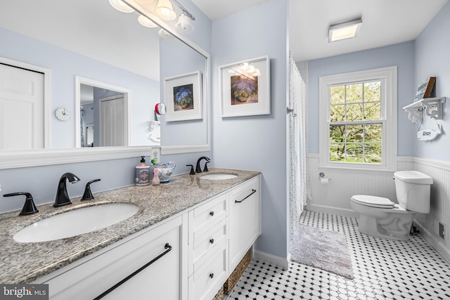 full bathroom with a sink, toilet, double vanity, and a wainscoted wall