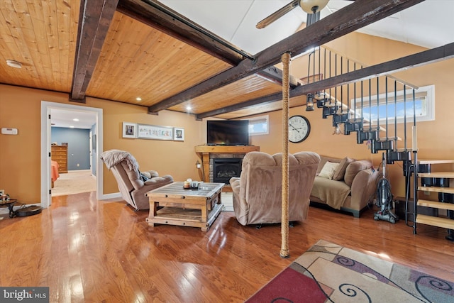 living area with ceiling fan, a stone fireplace, beam ceiling, wood finished floors, and stairs