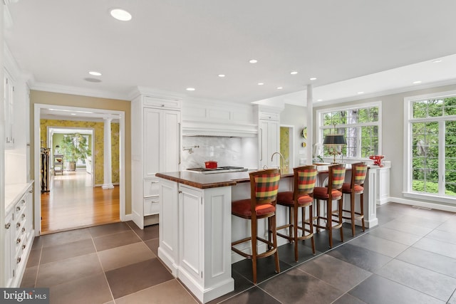 kitchen with an island with sink, dark tile patterned flooring, white cabinets, baseboards, and crown molding