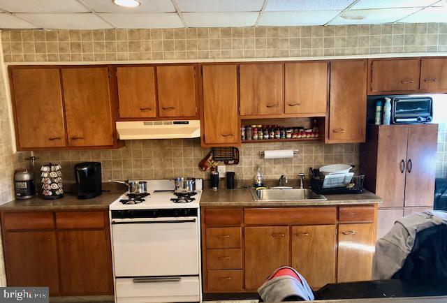 kitchen featuring backsplash, white gas range, under cabinet range hood, a drop ceiling, and a sink