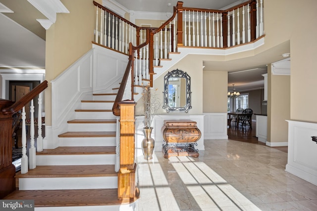 staircase featuring a notable chandelier, ornamental molding, a high ceiling, wainscoting, and a decorative wall