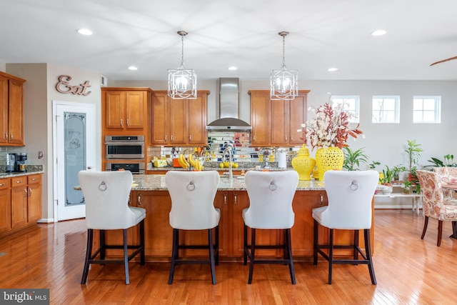 kitchen featuring light wood-style floors, wall chimney exhaust hood, brown cabinets, stainless steel double oven, and backsplash