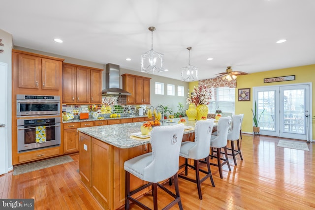 kitchen with wall chimney range hood, brown cabinets, recessed lighting, light stone countertops, and light wood-style flooring