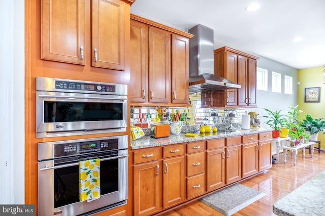 kitchen featuring wall chimney range hood, double oven, black electric stovetop, light wood-style flooring, and light stone counters