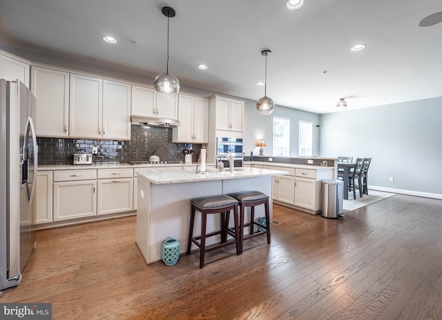 kitchen with a kitchen breakfast bar, backsplash, under cabinet range hood, hardwood / wood-style floors, and appliances with stainless steel finishes