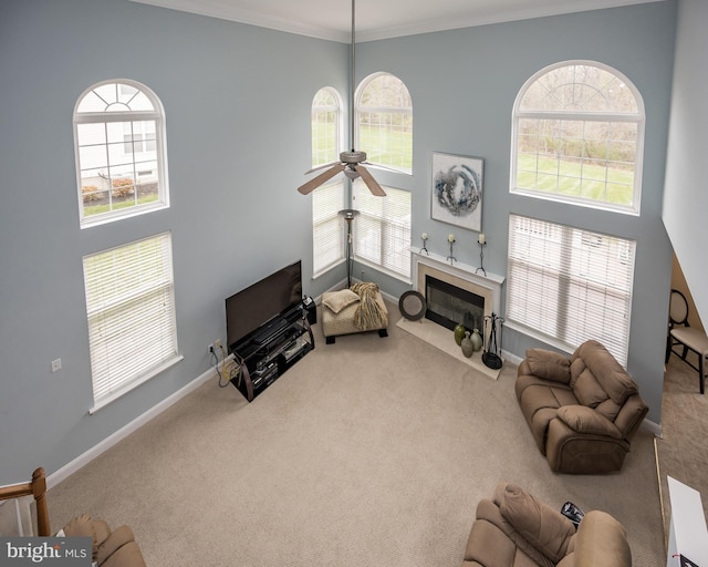 carpeted living area with baseboards, a high ceiling, a ceiling fan, crown molding, and a fireplace with flush hearth
