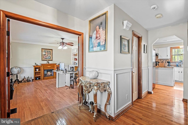 hall featuring built in shelves, arched walkways, ornamental molding, light wood-style floors, and wainscoting