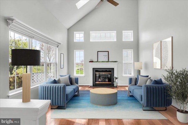 living area with high vaulted ceiling, a glass covered fireplace, a ceiling fan, and wood finished floors