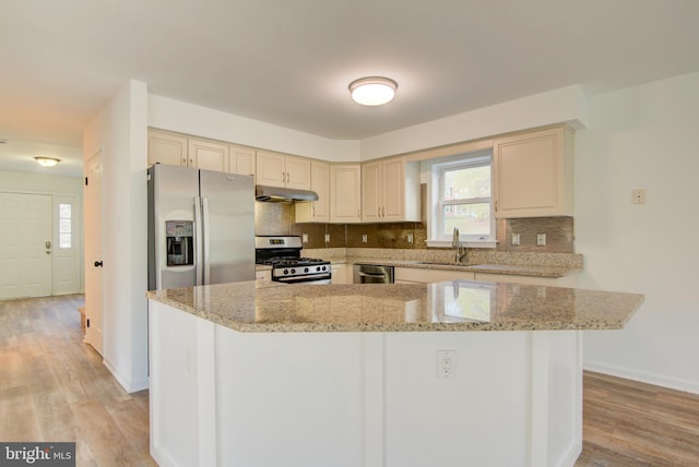 kitchen featuring cream cabinetry, under cabinet range hood, a sink, stainless steel appliances, and backsplash