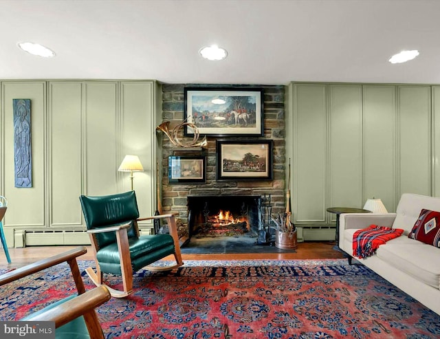 living area featuring a decorative wall, a stone fireplace, recessed lighting, wood finished floors, and a baseboard radiator
