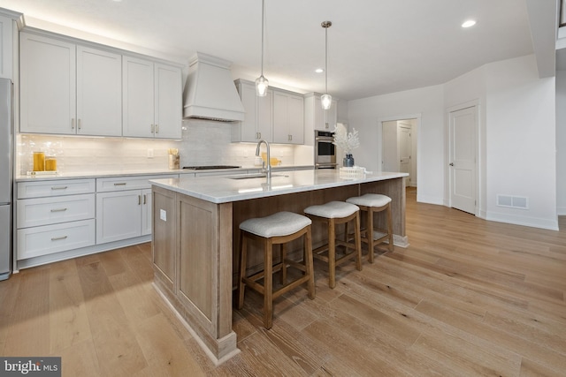 kitchen with visible vents, a sink, custom range hood, backsplash, and light wood-type flooring
