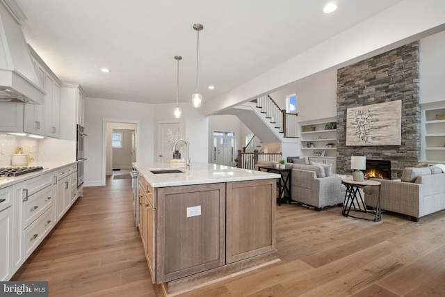 kitchen featuring an island with sink, a sink, light wood-type flooring, a stone fireplace, and custom range hood