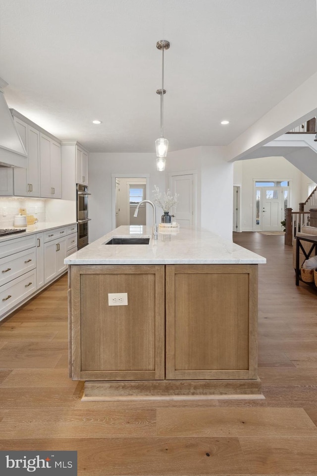 kitchen featuring an island with sink, light stone counters, a sink, double oven, and light wood-type flooring