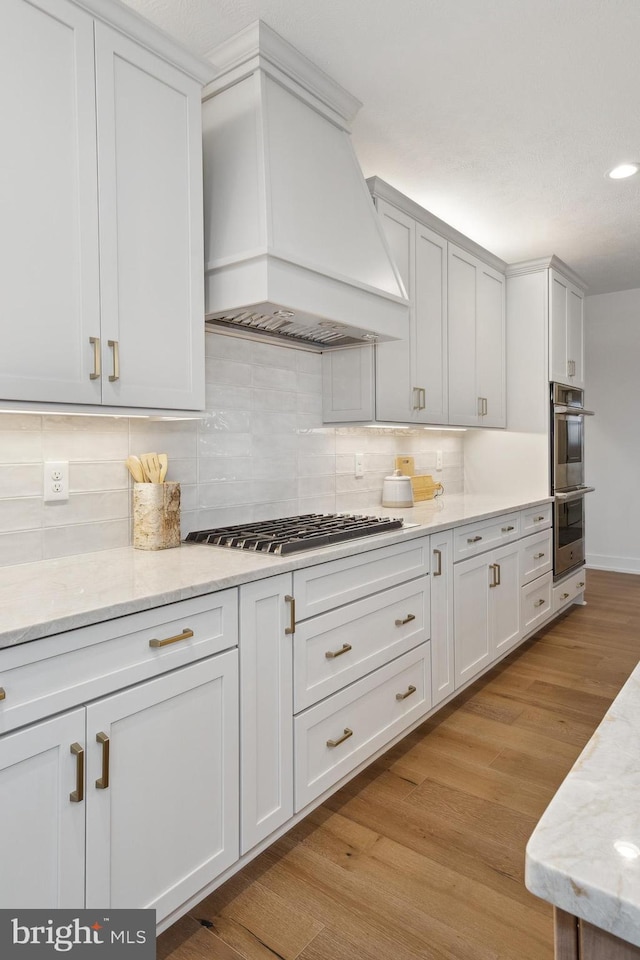 kitchen with custom range hood, white cabinetry, light wood-style floors, tasteful backsplash, and appliances with stainless steel finishes
