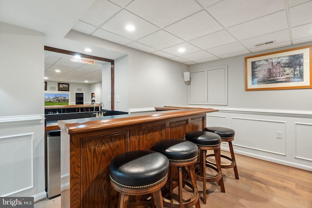 bar with light wood-style floors, a paneled ceiling, a decorative wall, a dry bar, and visible vents