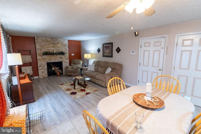 dining area featuring light wood-style floors, a stone fireplace, a textured ceiling, and ceiling fan