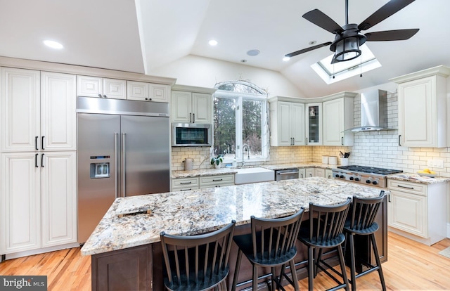 kitchen with a sink, lofted ceiling with skylight, a ceiling fan, wall chimney range hood, and appliances with stainless steel finishes