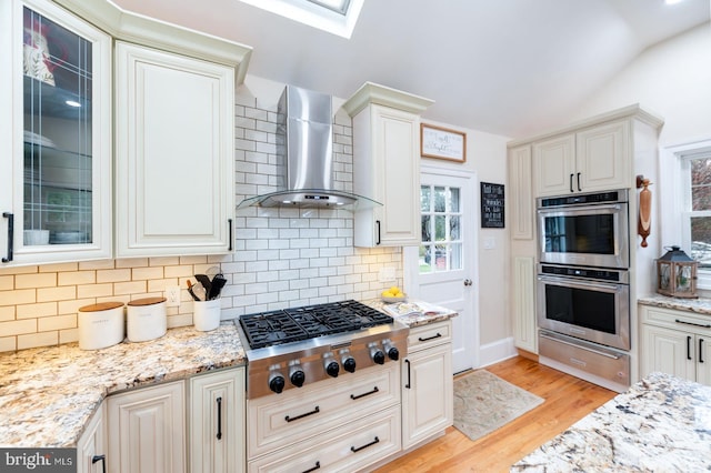 kitchen featuring wall chimney range hood, a warming drawer, stainless steel appliances, light wood finished floors, and backsplash