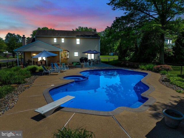 pool at dusk with a patio area, fence, a diving board, and a pool with connected hot tub