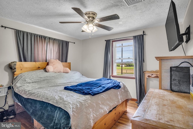 bedroom with ceiling fan, a textured ceiling, visible vents, a fireplace, and wood finished floors