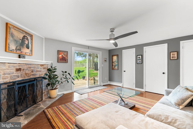 living area featuring wood finished floors, baseboard heating, ceiling fan, a fireplace, and baseboards