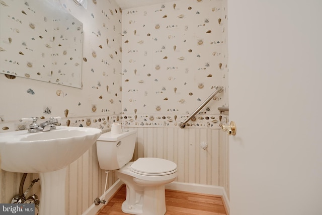 bathroom with wallpapered walls, wood finished floors, toilet, a sink, and baseboards