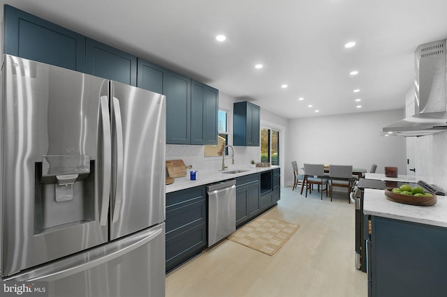 kitchen with a sink, wall chimney range hood, appliances with stainless steel finishes, backsplash, and blue cabinetry
