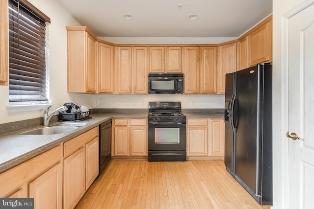 kitchen with light brown cabinets, a sink, dark countertops, black appliances, and light wood-style flooring