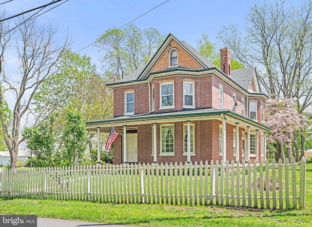 victorian-style house featuring a porch, a chimney, brick siding, and a fenced front yard