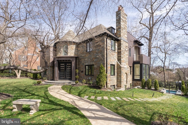 tudor home with stone siding, a chimney, fence, a balcony, and a front lawn