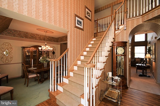 staircase with baseboards, arched walkways, an inviting chandelier, wood finished floors, and wallpapered walls