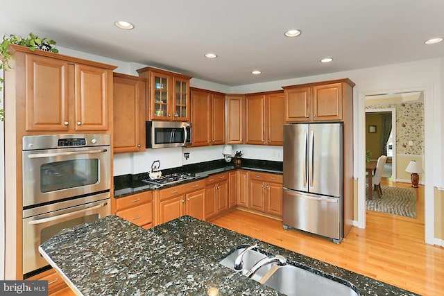 kitchen featuring dark stone counters, glass insert cabinets, a sink, appliances with stainless steel finishes, and light wood-type flooring