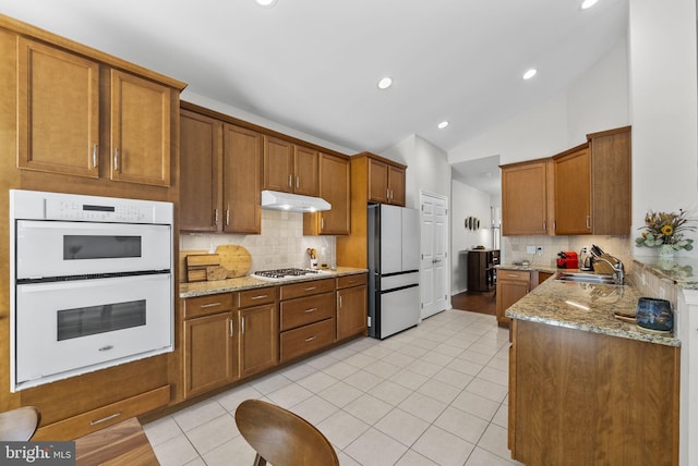 kitchen with white appliances, light tile patterned floors, under cabinet range hood, brown cabinetry, and a sink