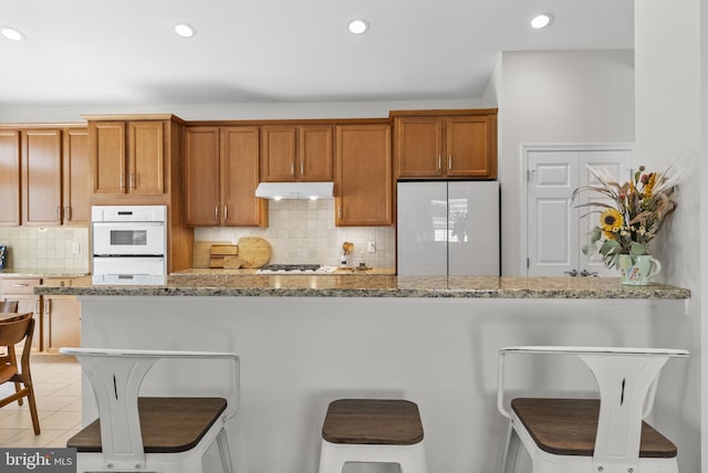 kitchen featuring under cabinet range hood, white appliances, a kitchen bar, and brown cabinets